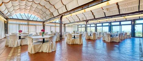 A spacious indoor venue with multiple round tables and chairs, large windows, and a wooden herringbone floor.