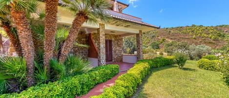 A house with a stone facade, a covered porch, and a well-maintained garden with palm trees and shrubs.