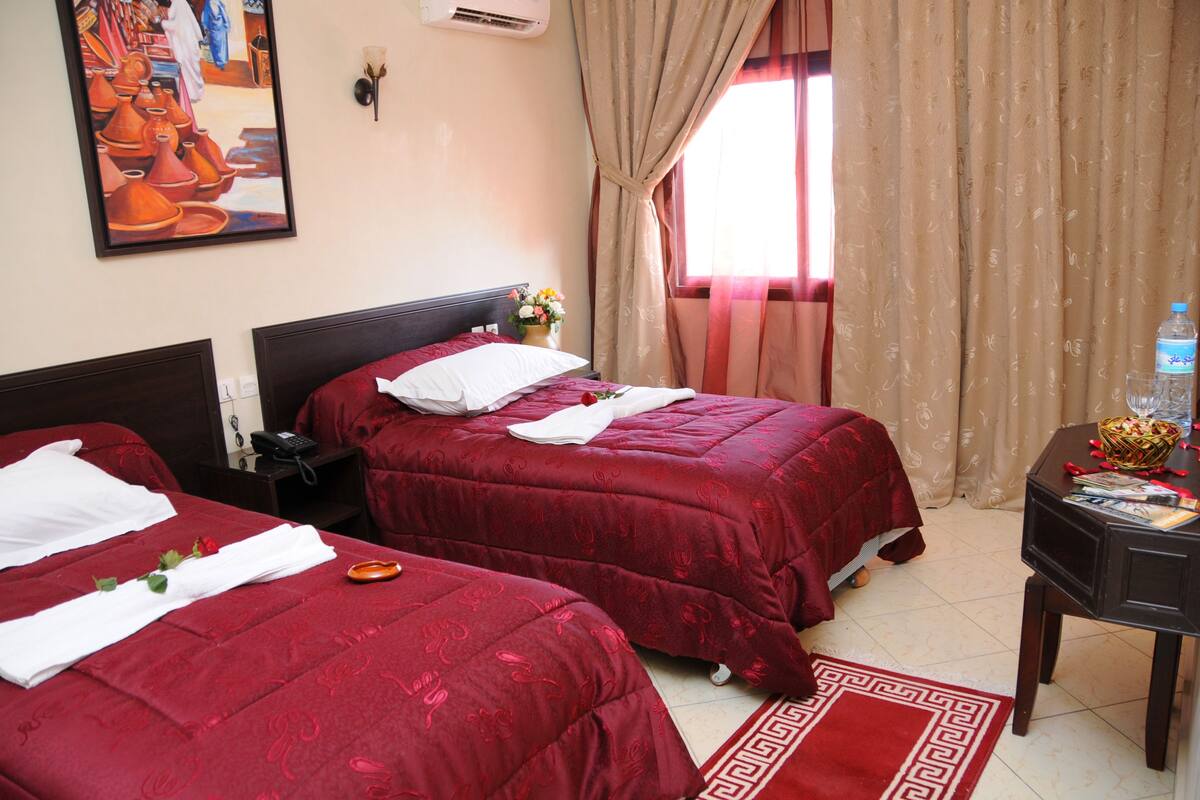 A hotel room with two beds, a nightstand, a table with a water bottle, and a painting on the wall.