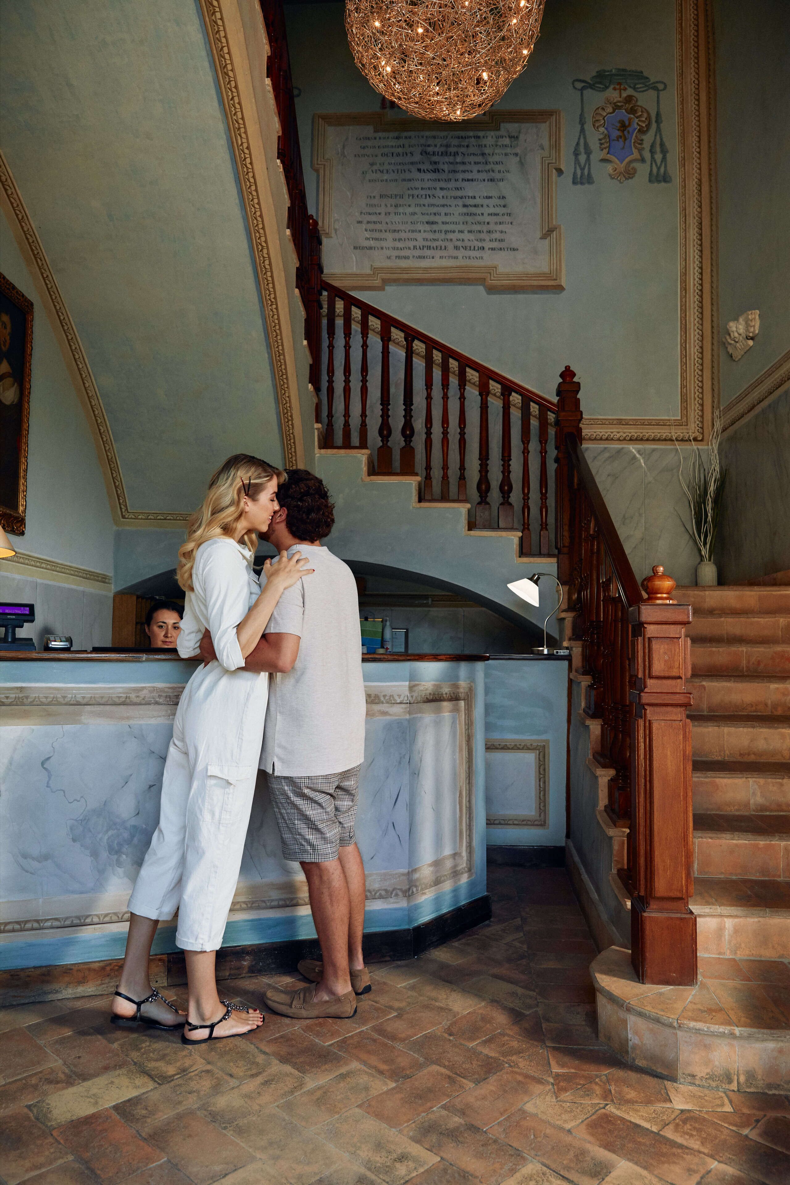A couple embracing in a grand hallway with a marble floor, a wooden staircase, and a chandelier.