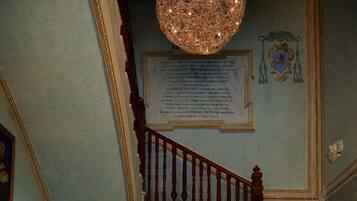 A couple embracing in a grand hallway with a marble floor, a wooden staircase, and a chandelier.
