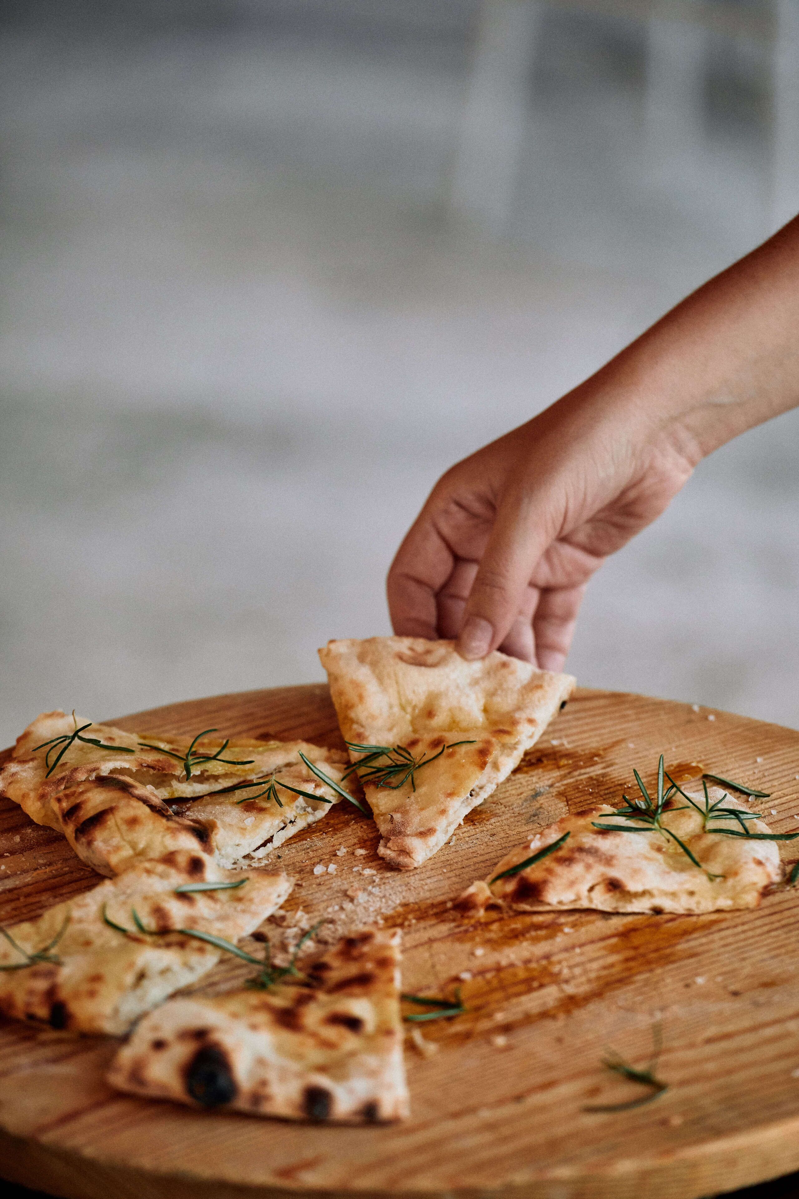 Una mano raccoglie un pezzo di pane piatto guarnito con erbe aromatiche da un tagliere di legno.