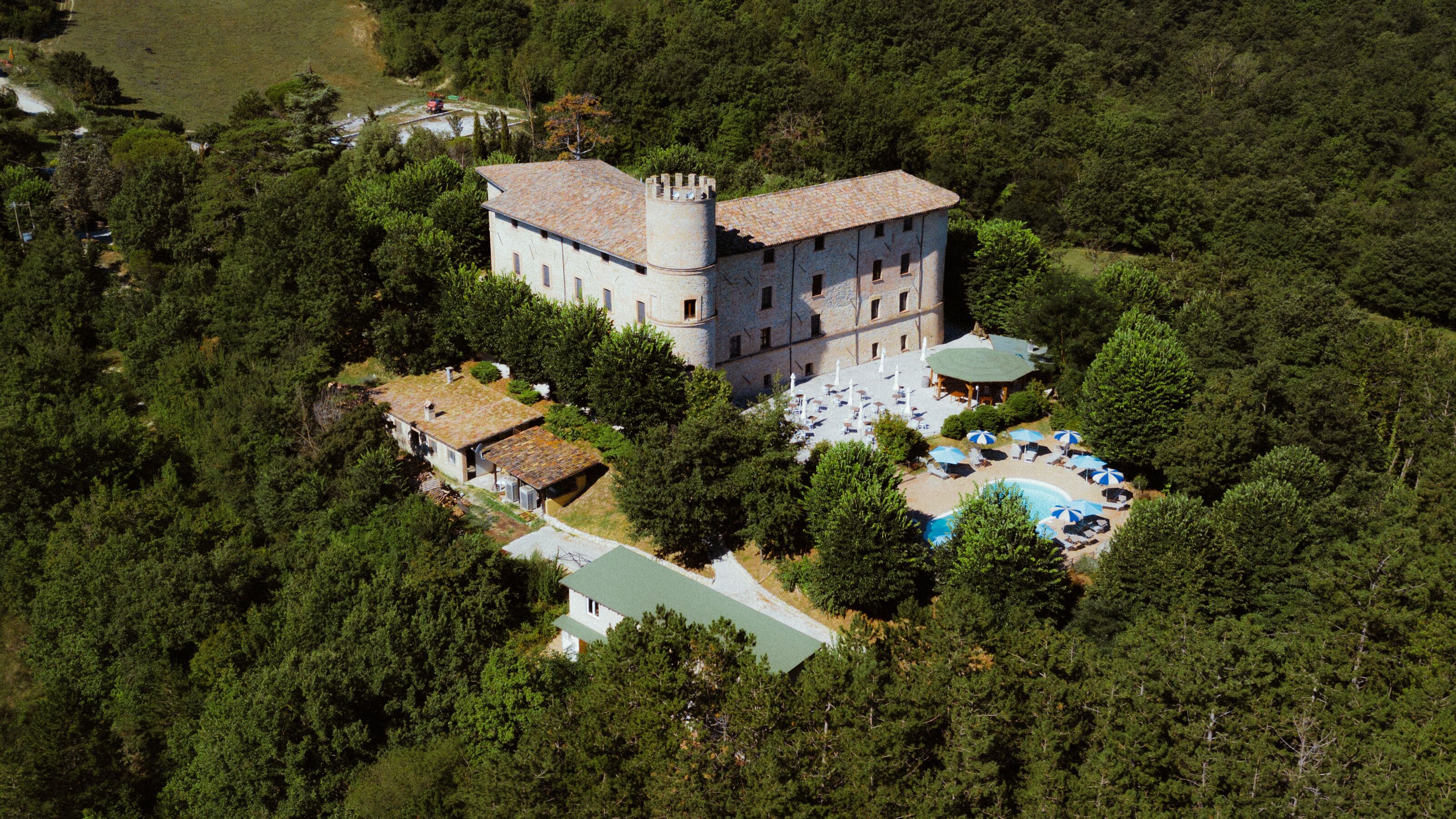 Une vue aérienne d’un bâtiment qui ressemble à un château, entouré d’une forêt dense, avec une piscine et un espace de détente extérieur.