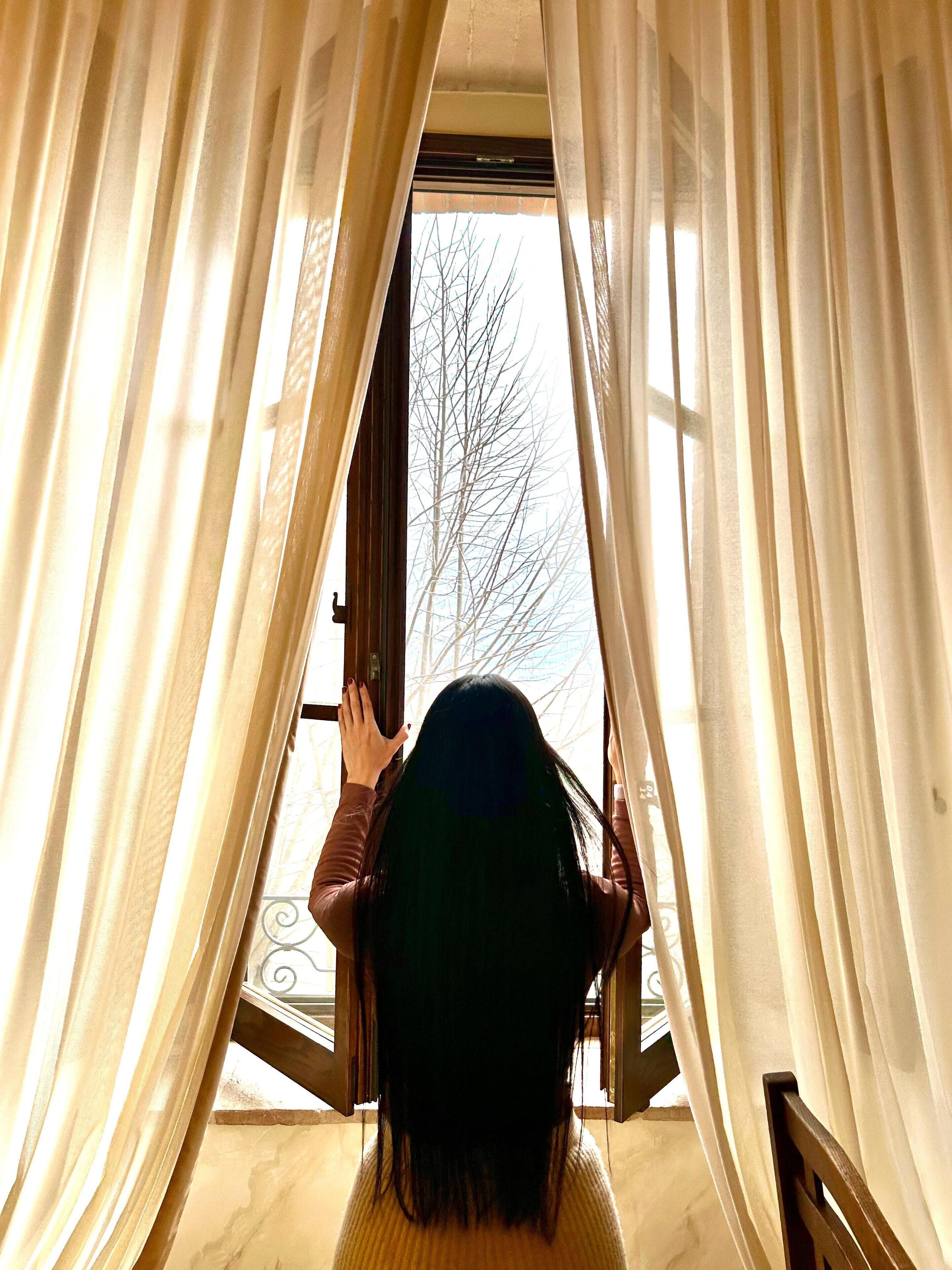 A person standing at a window with sheer curtains, looking outside.
