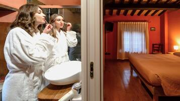A room with a bed, a wooden floor, a bathroom with a sink, and a mirror.