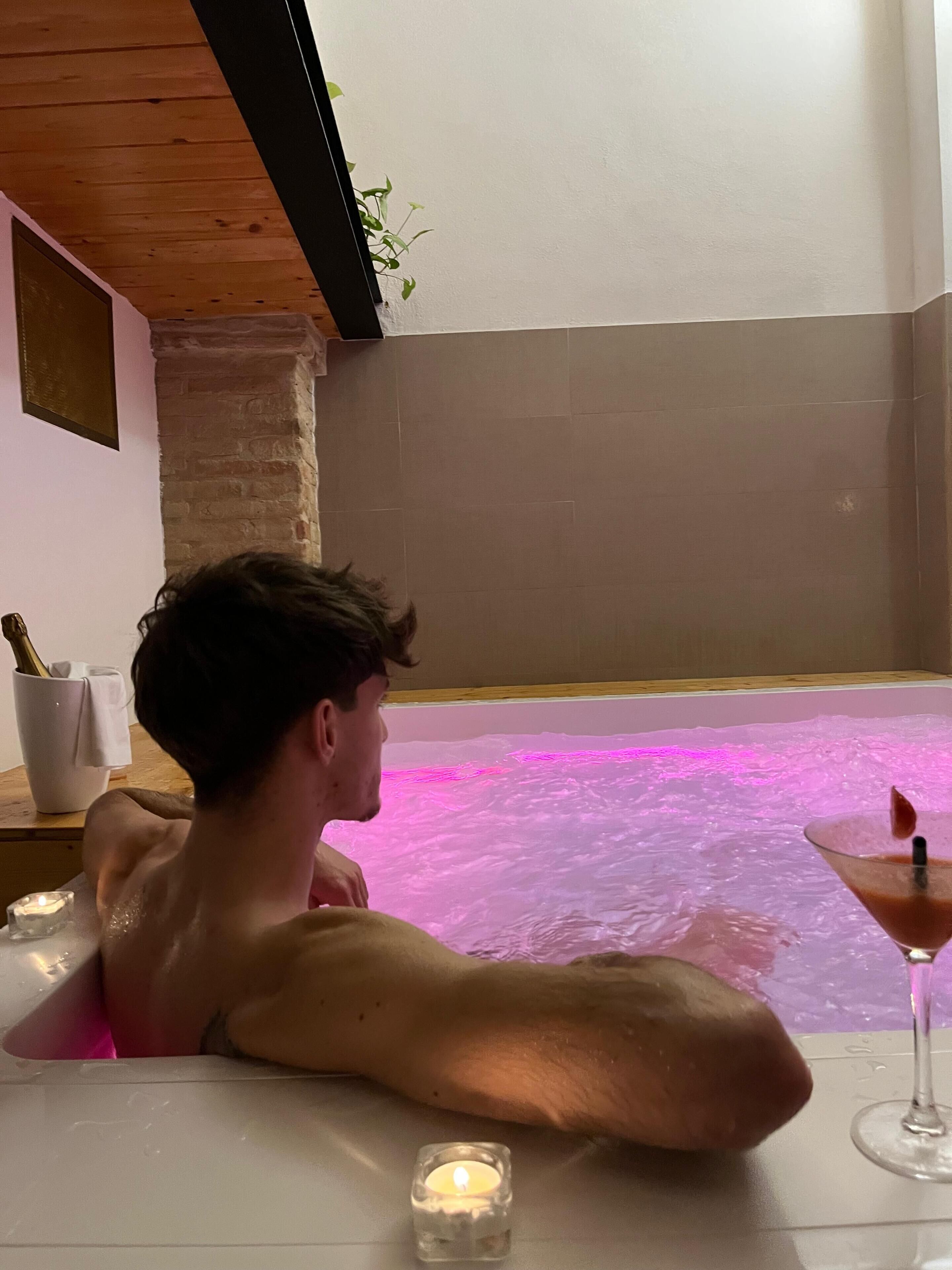 Iemand ontspant in een bubbelbad met roze licht, een kaars en een martini op een bijzettafeltje.