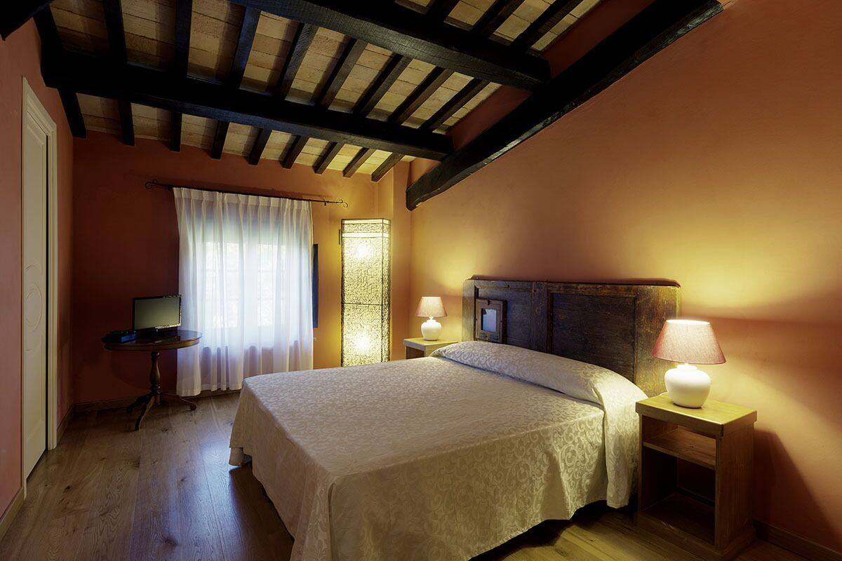 A bedroom with a bed, two bedside tables with lamps, a desk with a computer, a window with curtains, and a wooden ceiling.