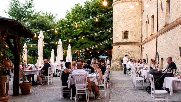 Un área de comedor al aire libre con mesas y sillas, luces colgantes y un edificio de piedra al fondo.