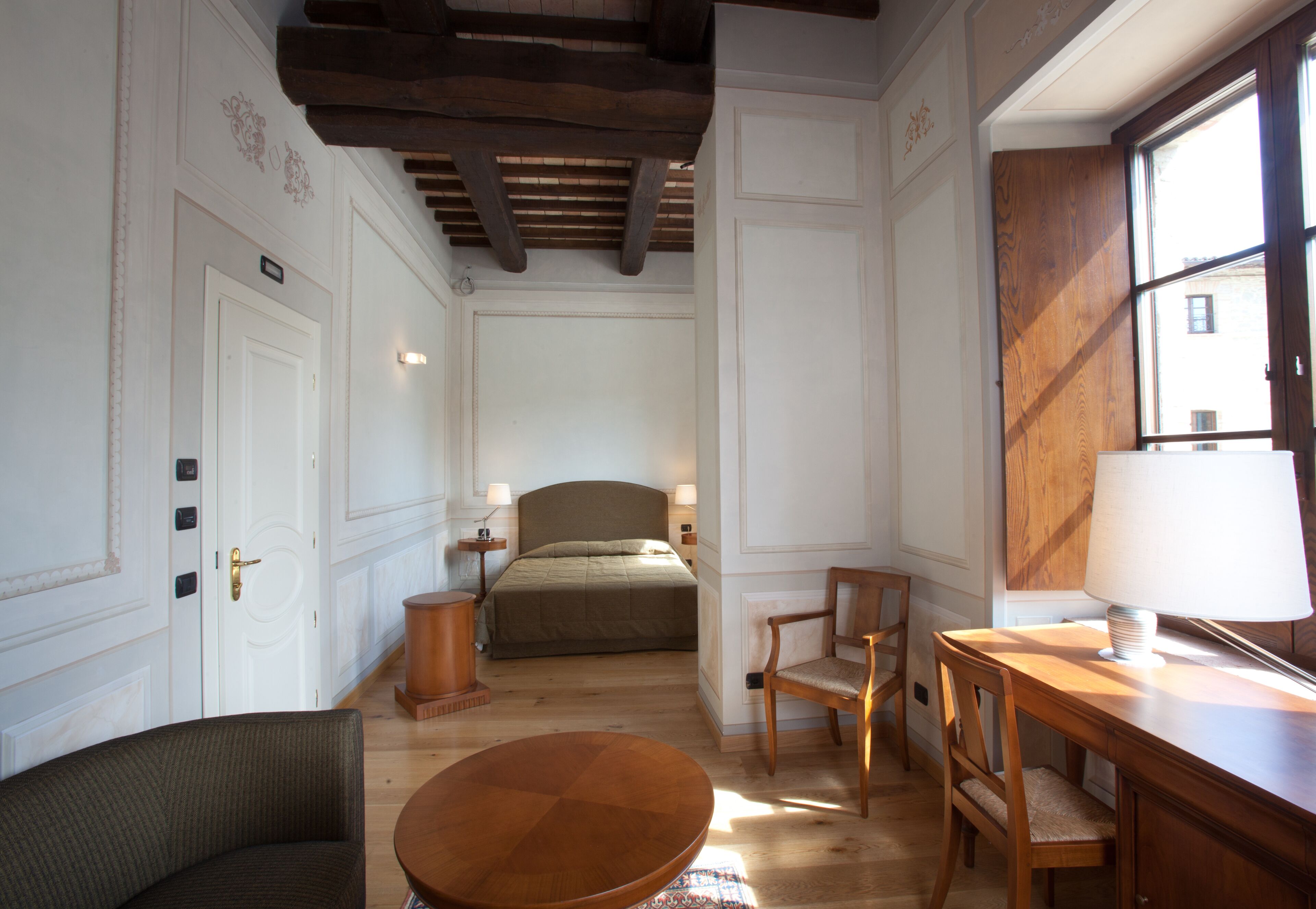 A room with a bed, a desk with a lamp, a chair, and a round table.