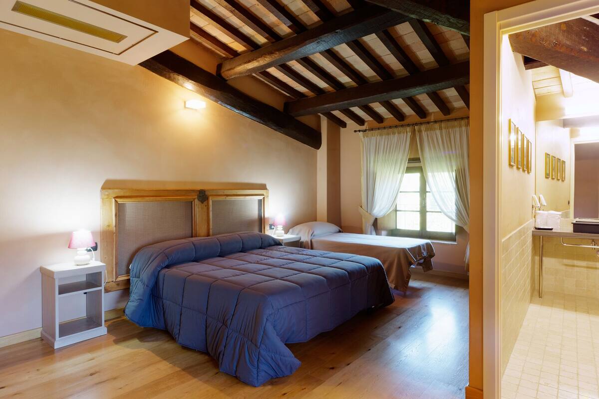A bedroom with a large bed, a bedside table, a window with curtains, and a wooden ceiling.