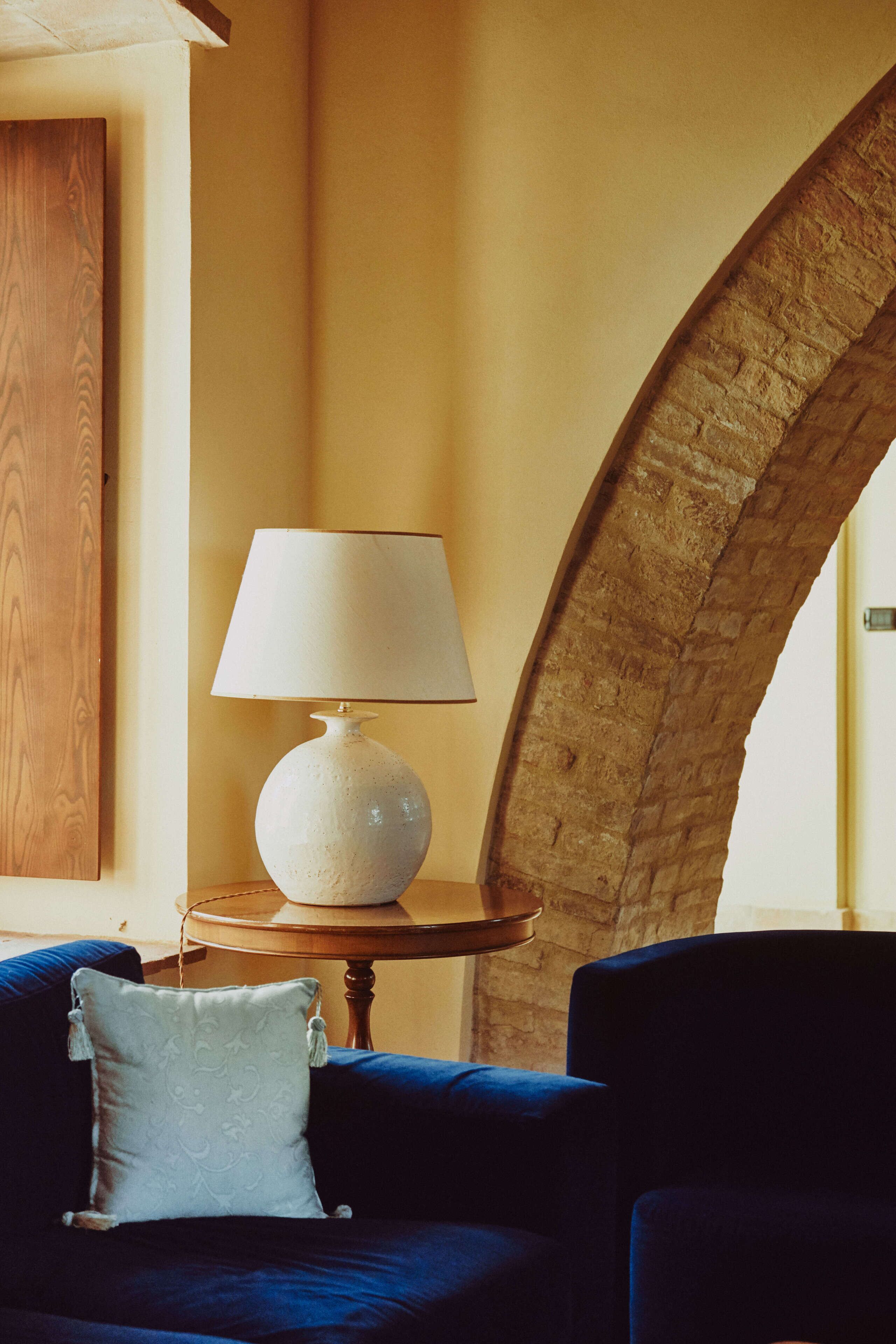 Una habitación con un sofá azul, una lámpara blanca sobre una mesa de madera y un arco de piedra.