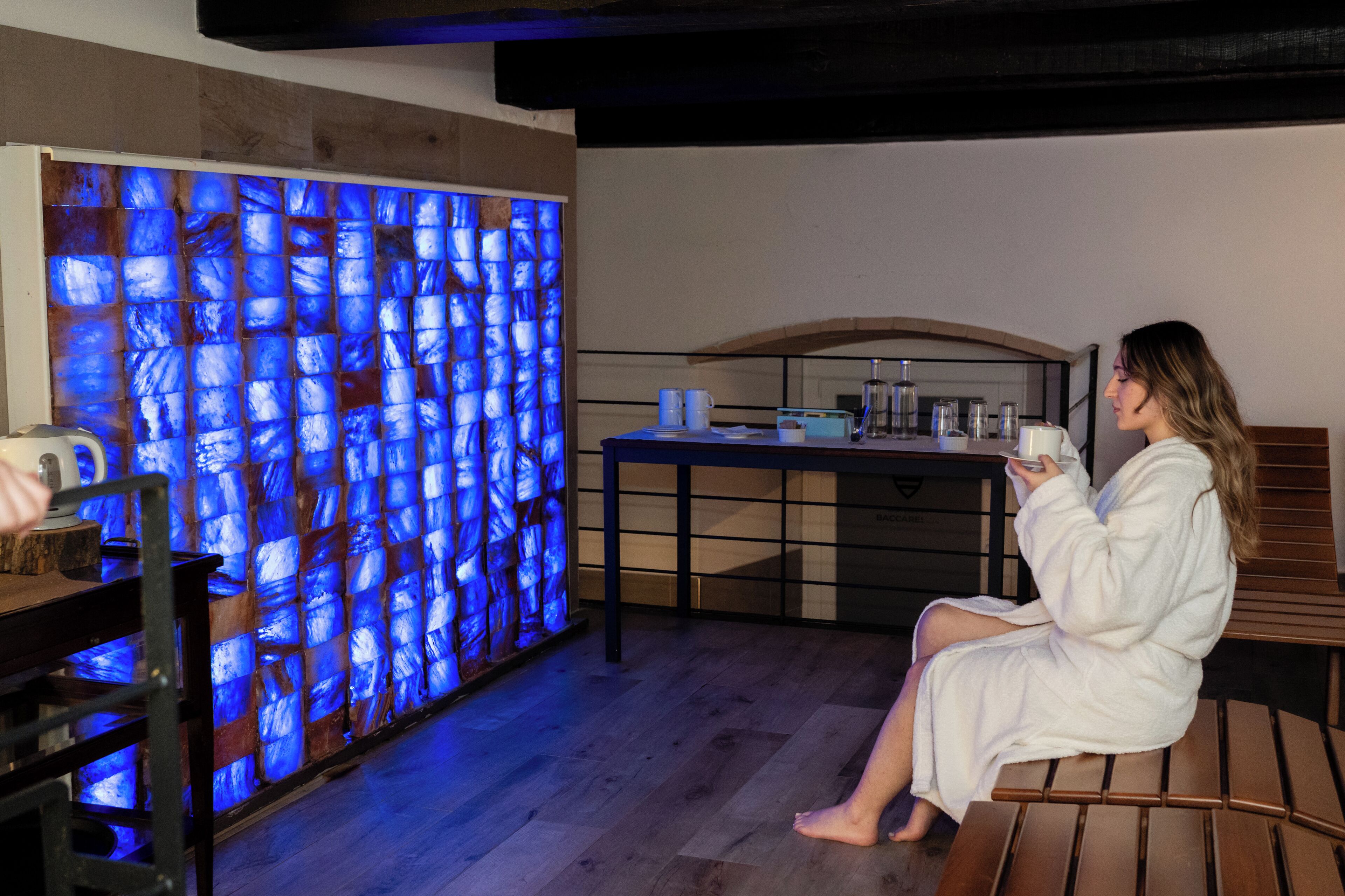 A person in a white bathrobe sitting on a wooden bench in a room with a blue glass wall and a table with various items.