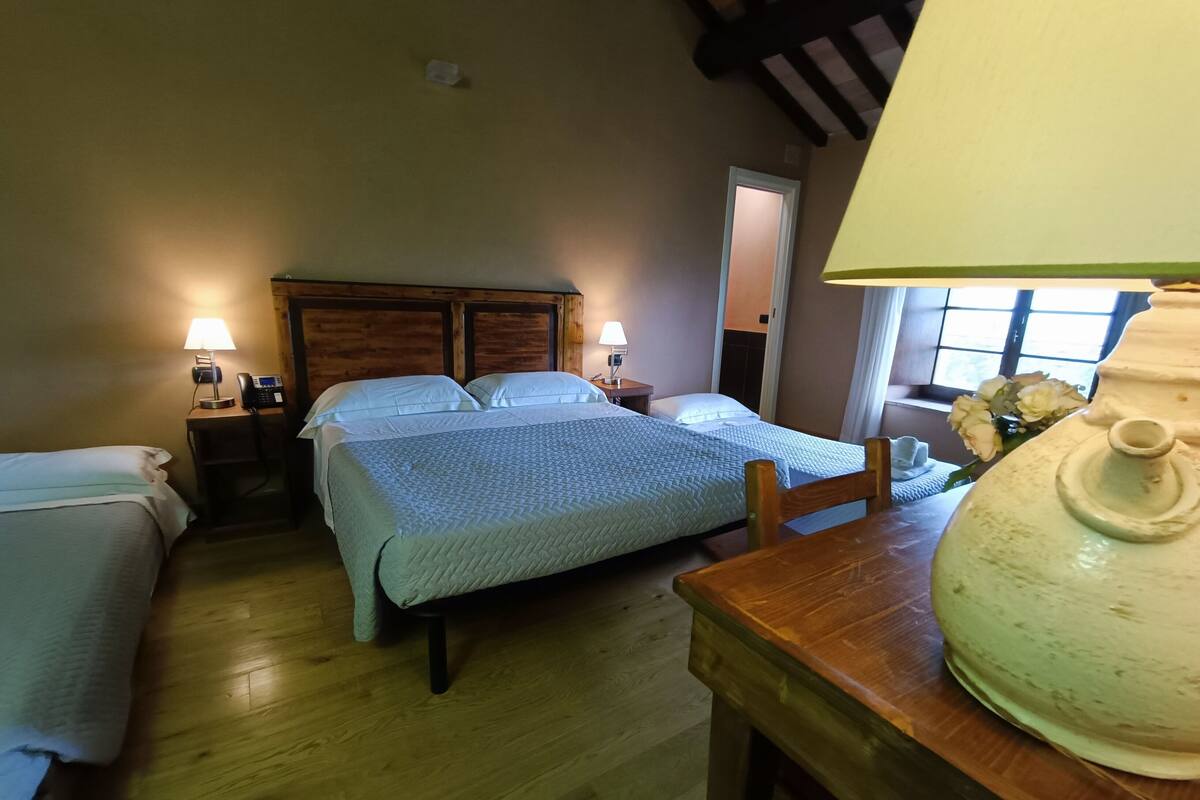 A cozy room with two beds, wooden flooring, a table with a lamp, and a window with flowers.