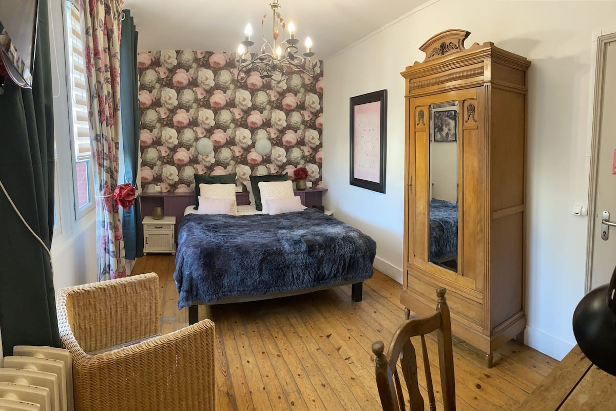 A bedroom with a bed, a dresser, a chair, a wooden floor, a floral wallpaper, and a chandelier.