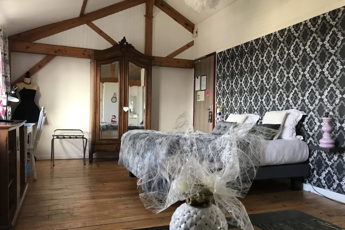 A bedroom with a bed, wooden beams, a dresser, a mirror, and patterned wallpaper.