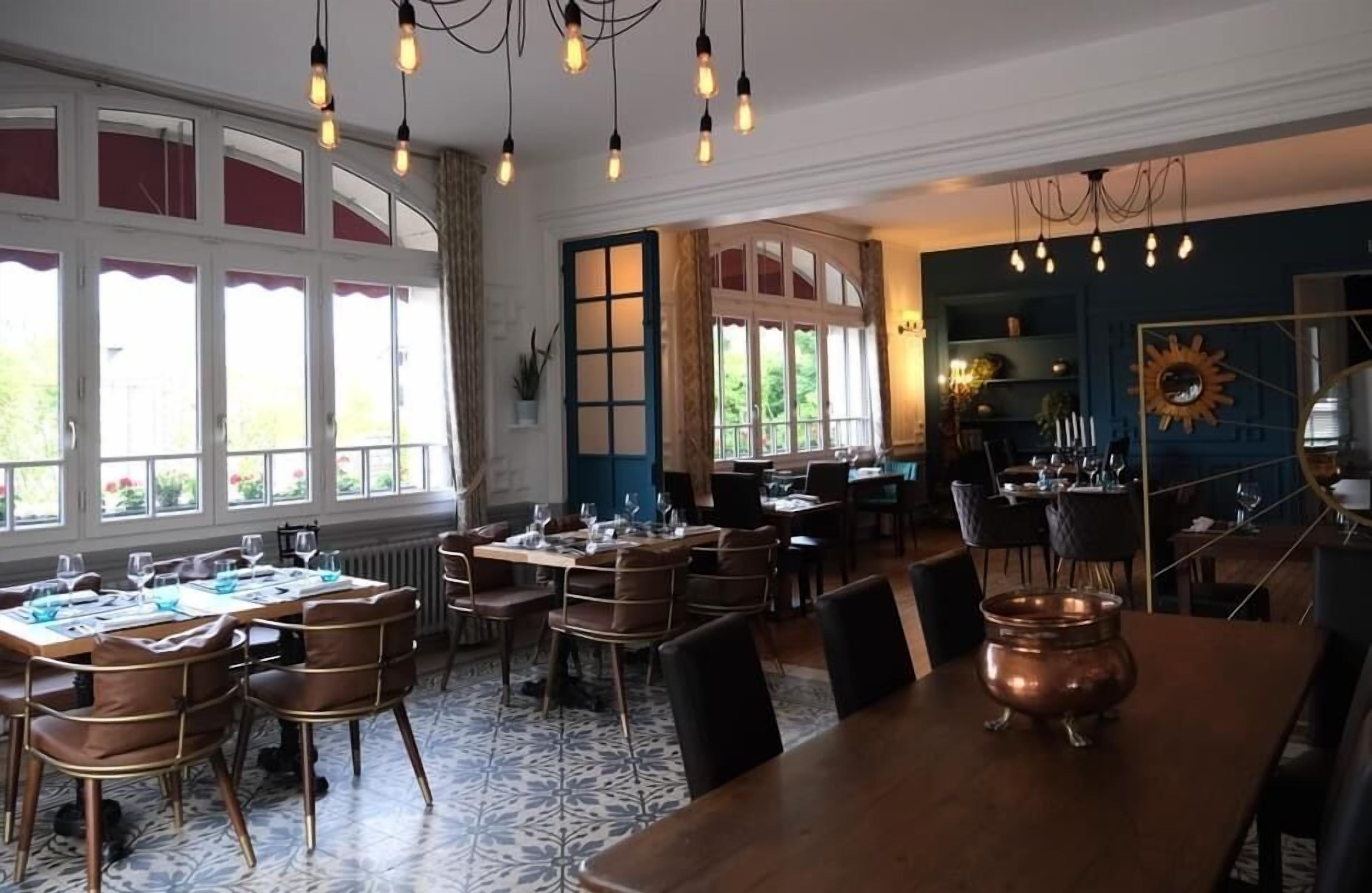 Un comedor con mesas dispuestas para una comida, ventanas amplias y una olla decorativa de cobre sobre la mesa.