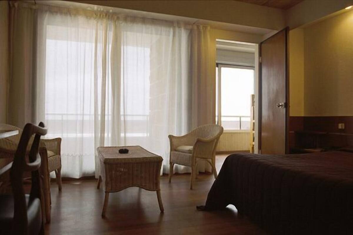 A room with a bed, a small table, two chairs, and a window with sheer curtains.