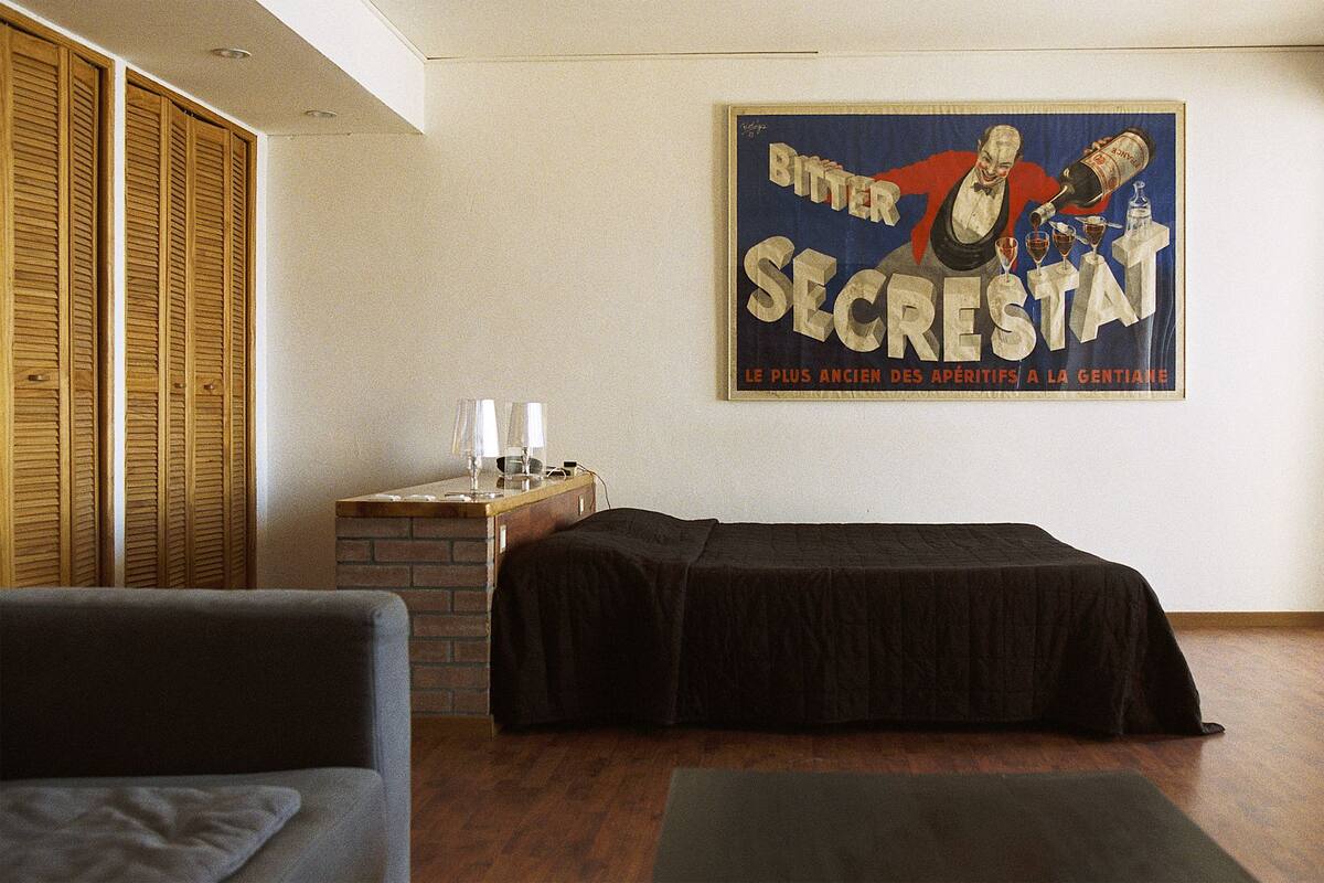 A hotel room with a bed, a small table with a lamp, and a poster on the wall.