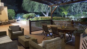 An outdoor patio area with wicker furniture, a wooden table, and a view of a landscaped garden at night. - B&B Villaseta (Agrigento)