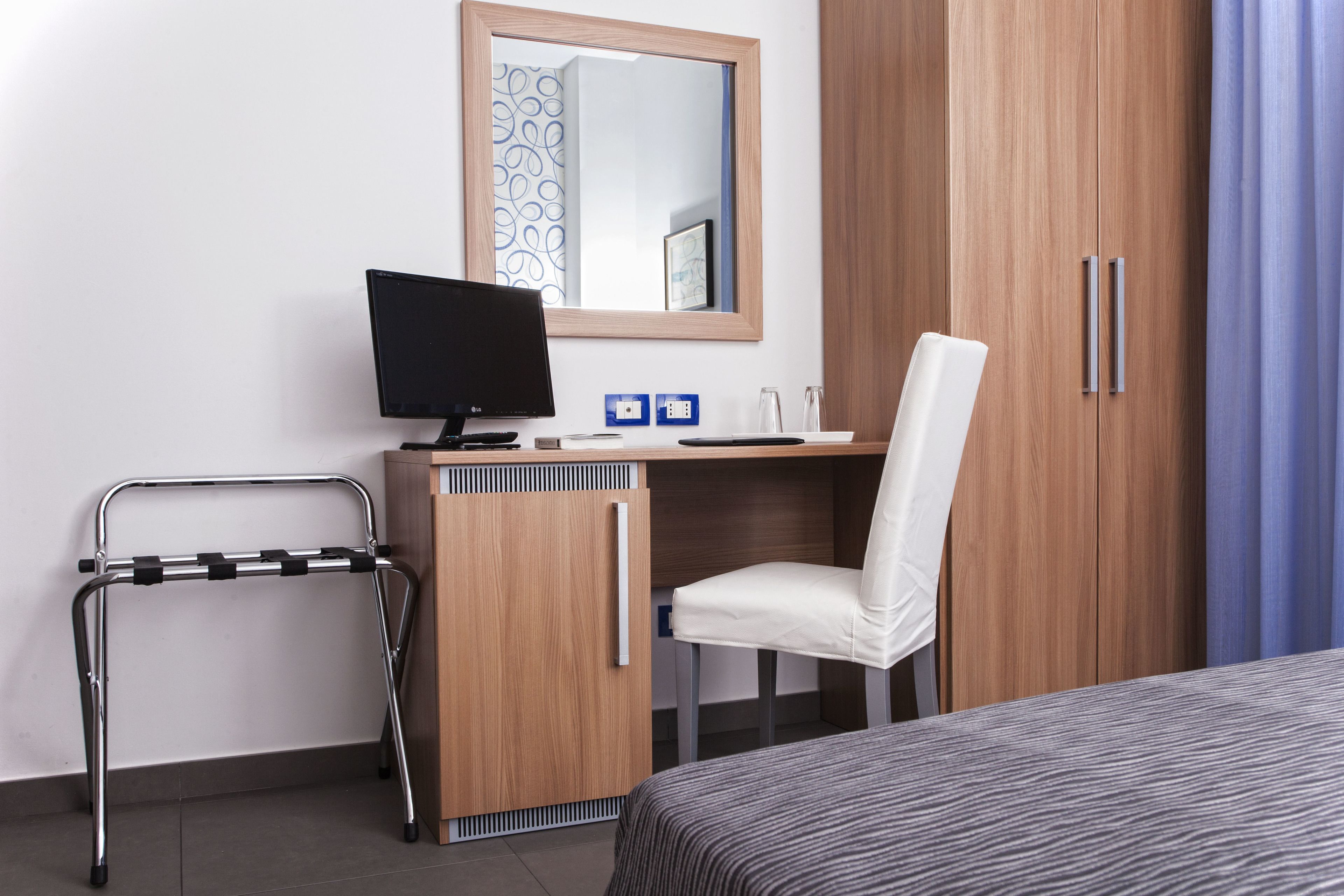 Une chambre d’hôtel avec un bureau en bois, un ordinateur, une chaise blanche, un lit et un miroir.
