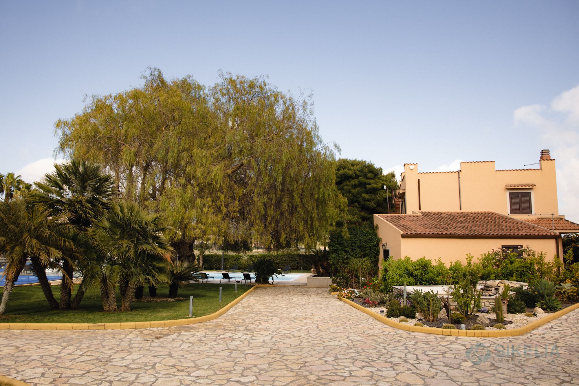 A paved pathway leading to a building with a tiled roof, surrounded by lush greenery and a variety of plants.