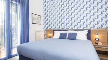 Een groot bed met een geborduurde blauwe sprei, twee nachtkastjes met lampen, een raam met blauwe gordijnen, en behang met een patroon.