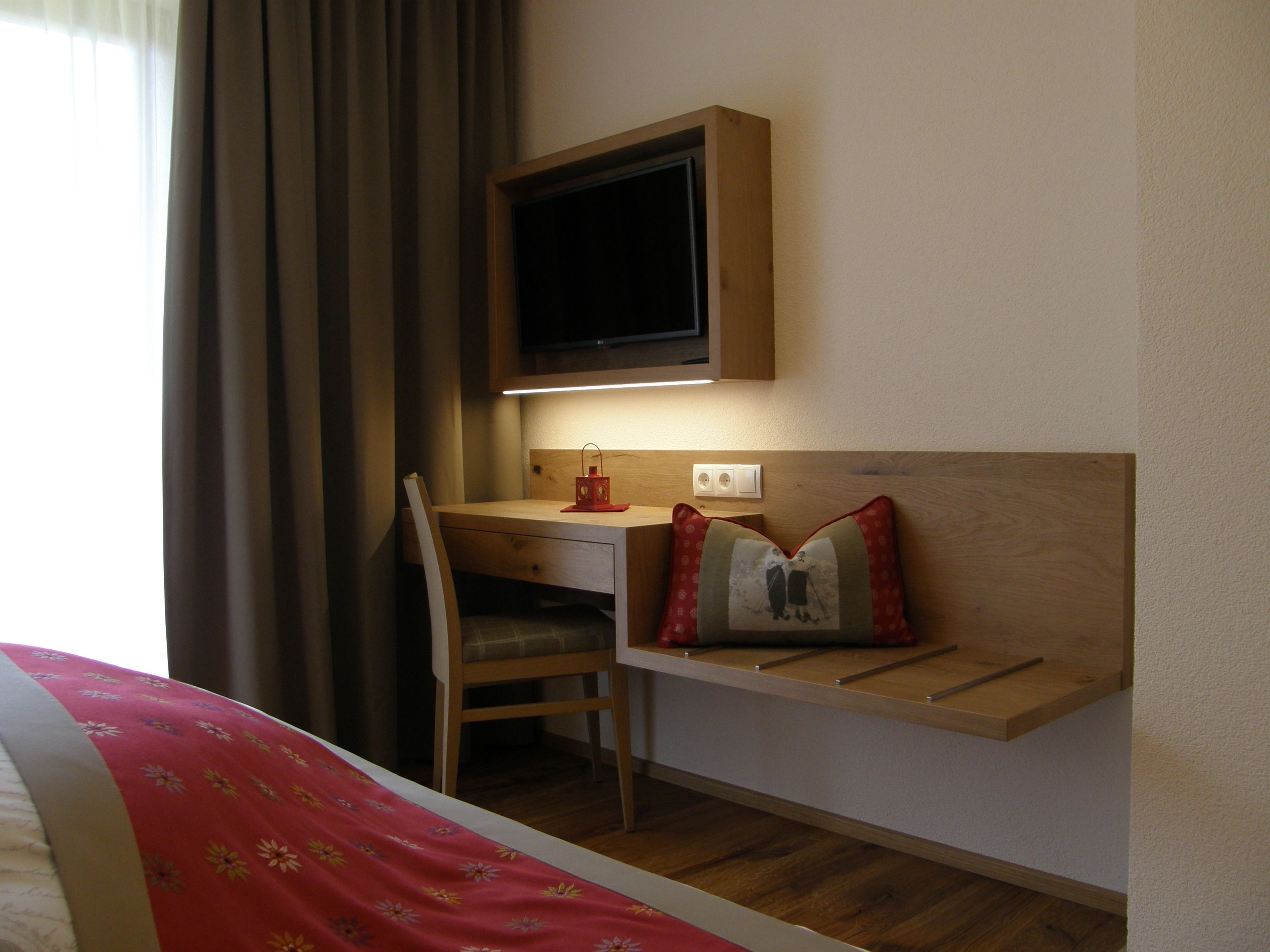 Habitación de hotel con escritorio de madera, una silla, una cama con colcha estampada de flores rojas, televisión montada en la pared y una mesita de noche con almohada roja y gris.