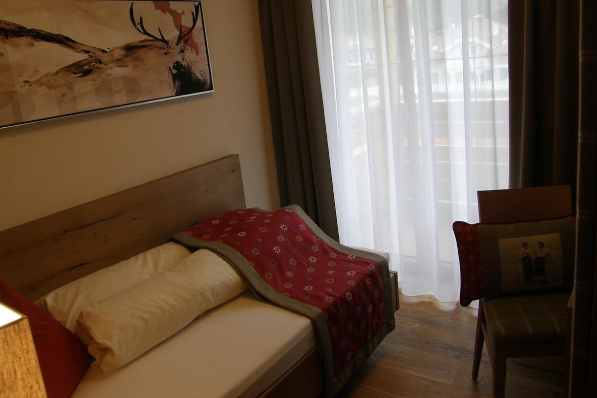 A hotel room with a bed, a chair, a window with curtains, and a wall-mounted painting.