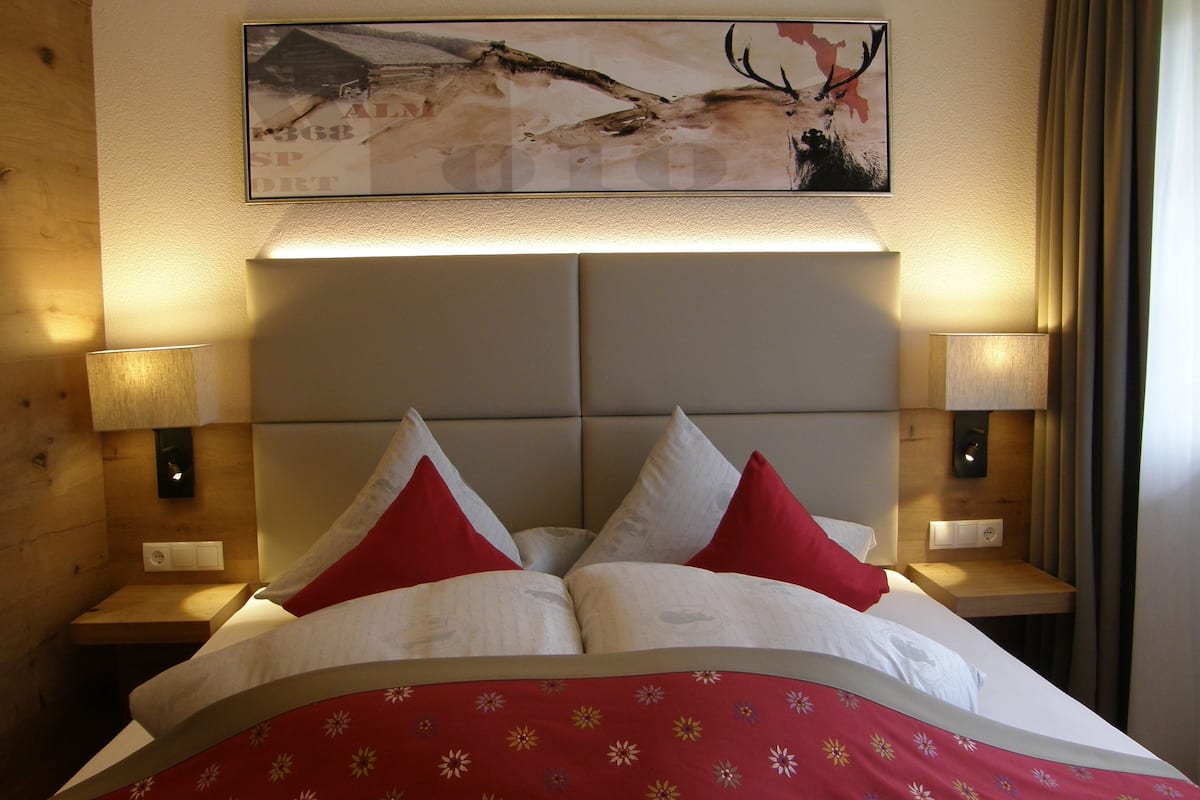 A hotel room with a large bed, two bedside lamps, a headboard with a mounted painting, and a wooden headboard on one side.