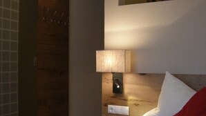 A bedroom with a wooden floor, a bed with white and red bedding, a nightstand with a lamp, a mirror on the wall, and a door with a towel rack. - Pension Sportalm (Soelden)