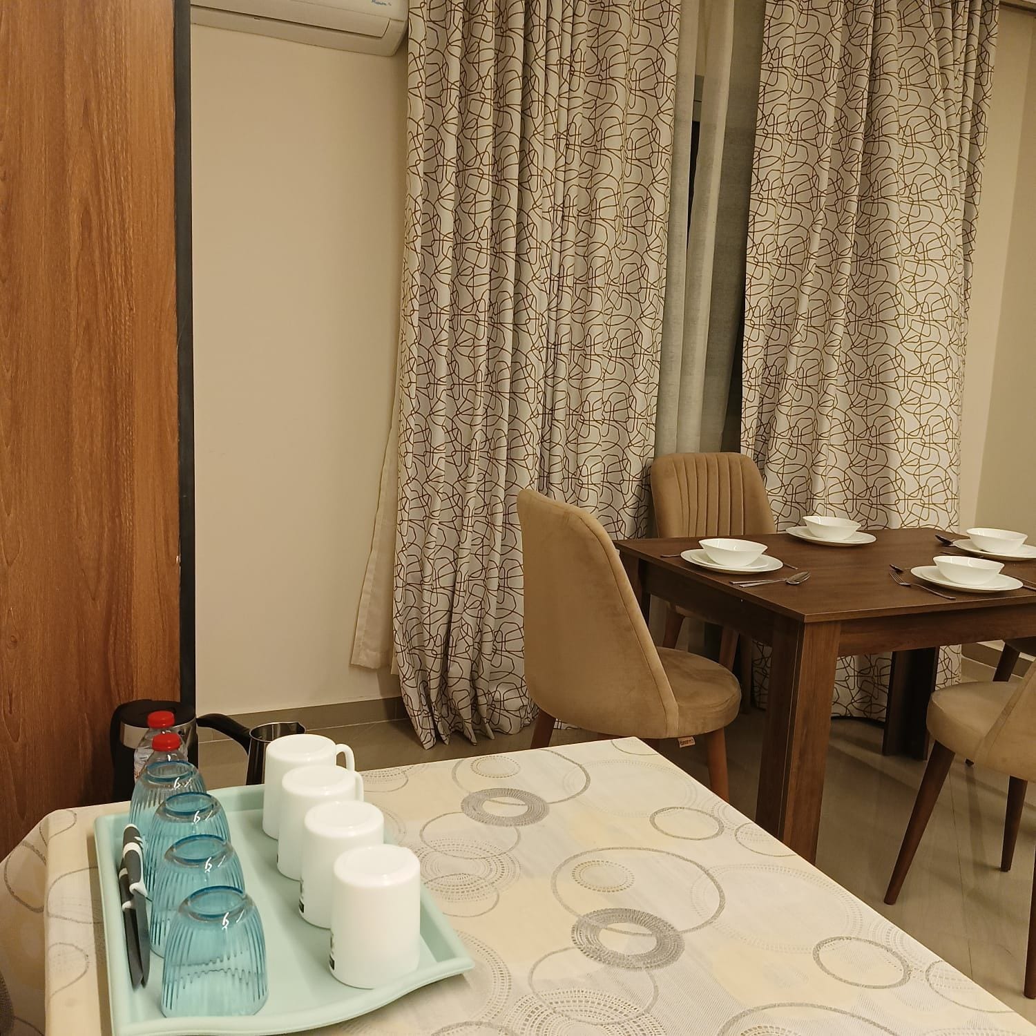 A dining area with a table set for two, a tray with water bottles and glasses, and curtains with a geometric pattern.