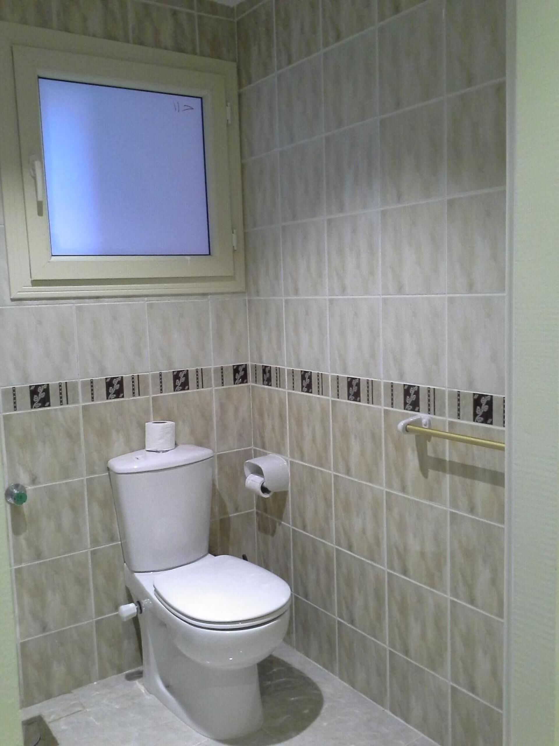 Un baño con un inodoro, un dispensador de papel higiénico y una ventana sobre el inodoro.