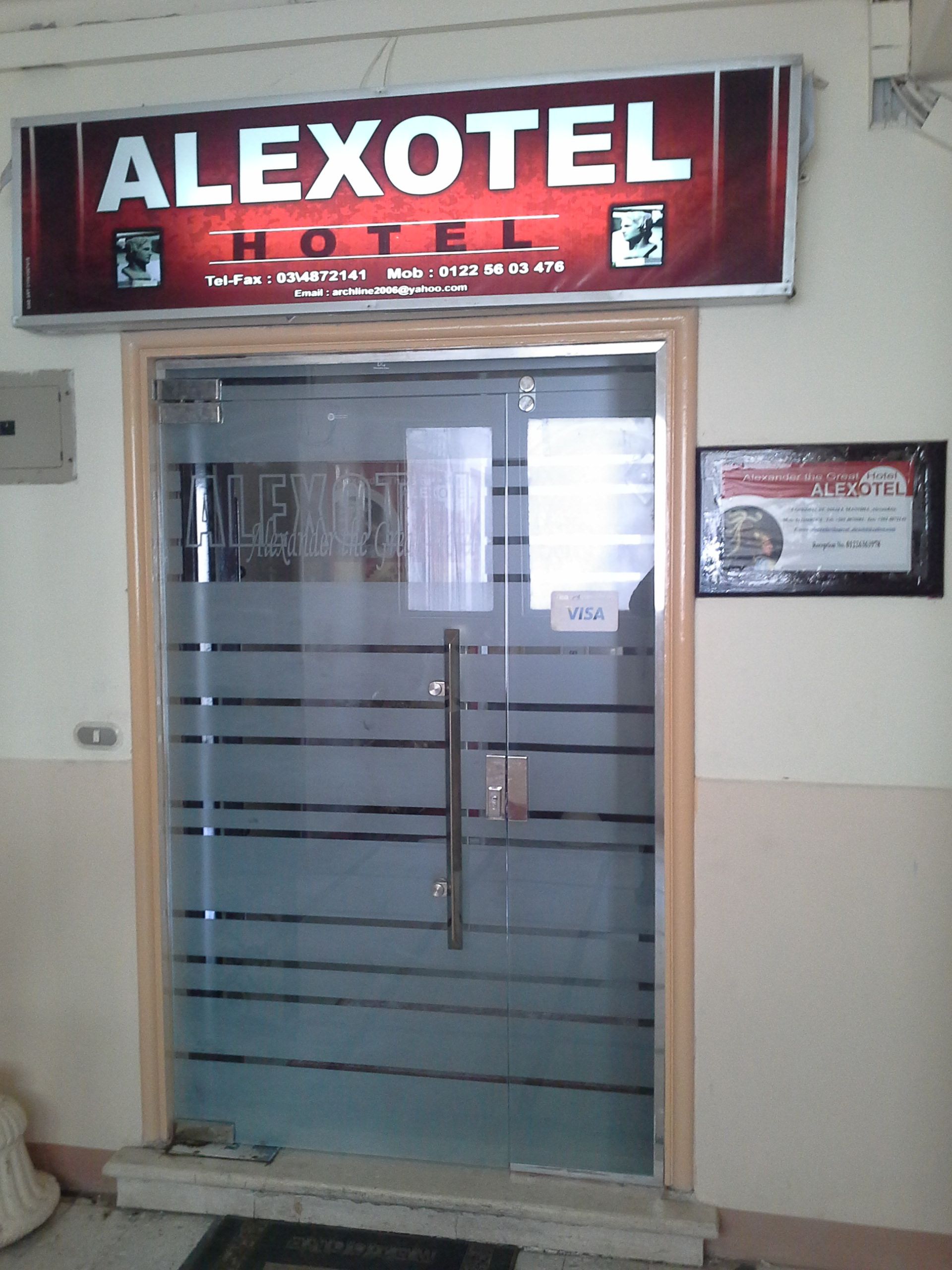 A hotel entrance with a glass door, a sign above stating "ALEXOTEL HOTEL", and a framed certificate on the wall.