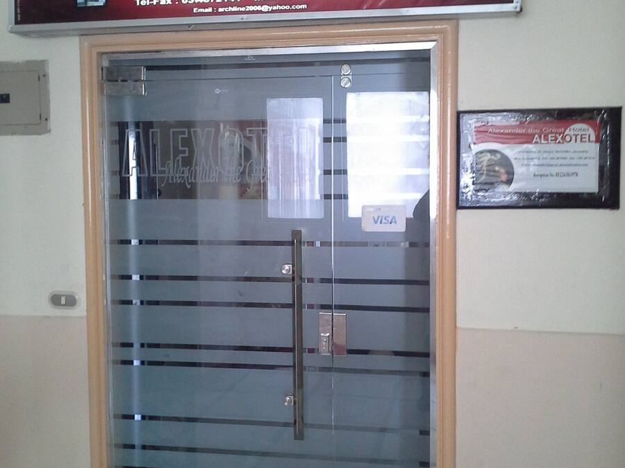 A hotel entrance with a glass door, a sign above stating "ALEXOTEL HOTEL", and a framed certificate on the wall.