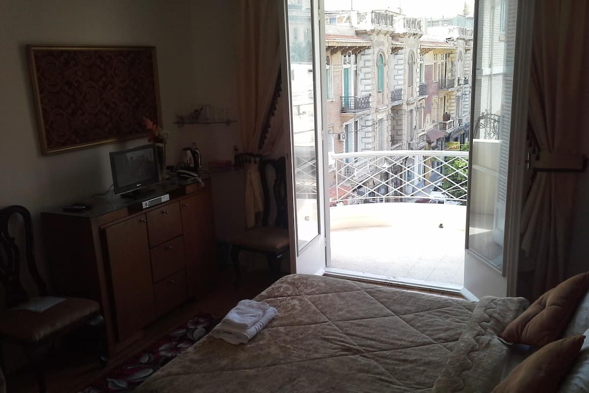 A hotel room with a bed, a desk with a computer, a chair, a window with a view of buildings, and a balcony with a railing.