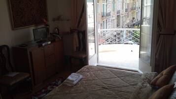 Una camera d'albergo con un letto, una scrivania con un computer, una sedia, una finestra con vista sugli edifici e un balcone con ringhiera.