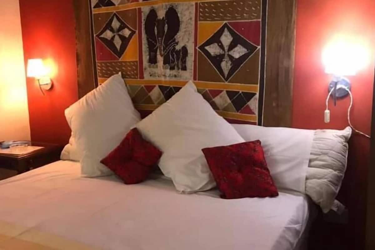 A bed with white linens and red pillows, a nightstand with a lamp, and a wall with a geometric patterned tapestry.