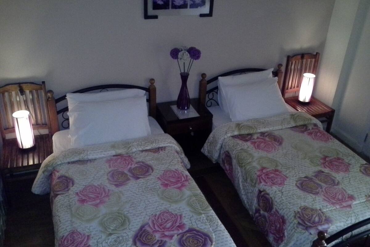 A room with two beds, each with a floral bedspread, a nightstand with a lamp, and a framed picture on the wall.