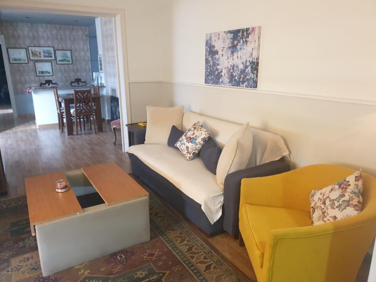A living room with a sofa, a yellow armchair, a coffee table, and a rug. There is a dining area in the background with a table and chairs.
