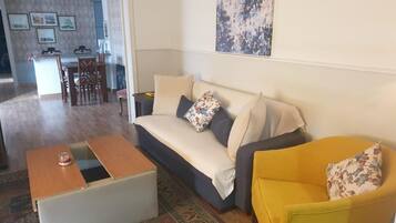 A living room with a sofa, a yellow armchair, a coffee table, and a rug. There is a dining area in the background with a table and chairs.
