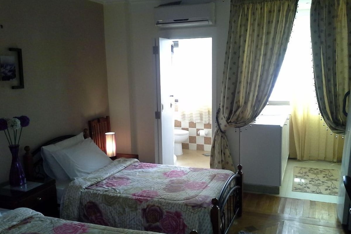 Habitación de hotel con dos camas, una mesita de noche, una lámpara, una ventana con cortinas y vistas al baño y al balcón.