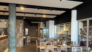 A modern restaurant interior with wooden tables and chairs, a bar area, and a view of the street through large windows. - Hotel Salgueiro (Porto Moniz)