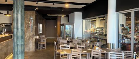 A modern restaurant interior with wooden tables and chairs, a bar area, and a view of the street through large windows.