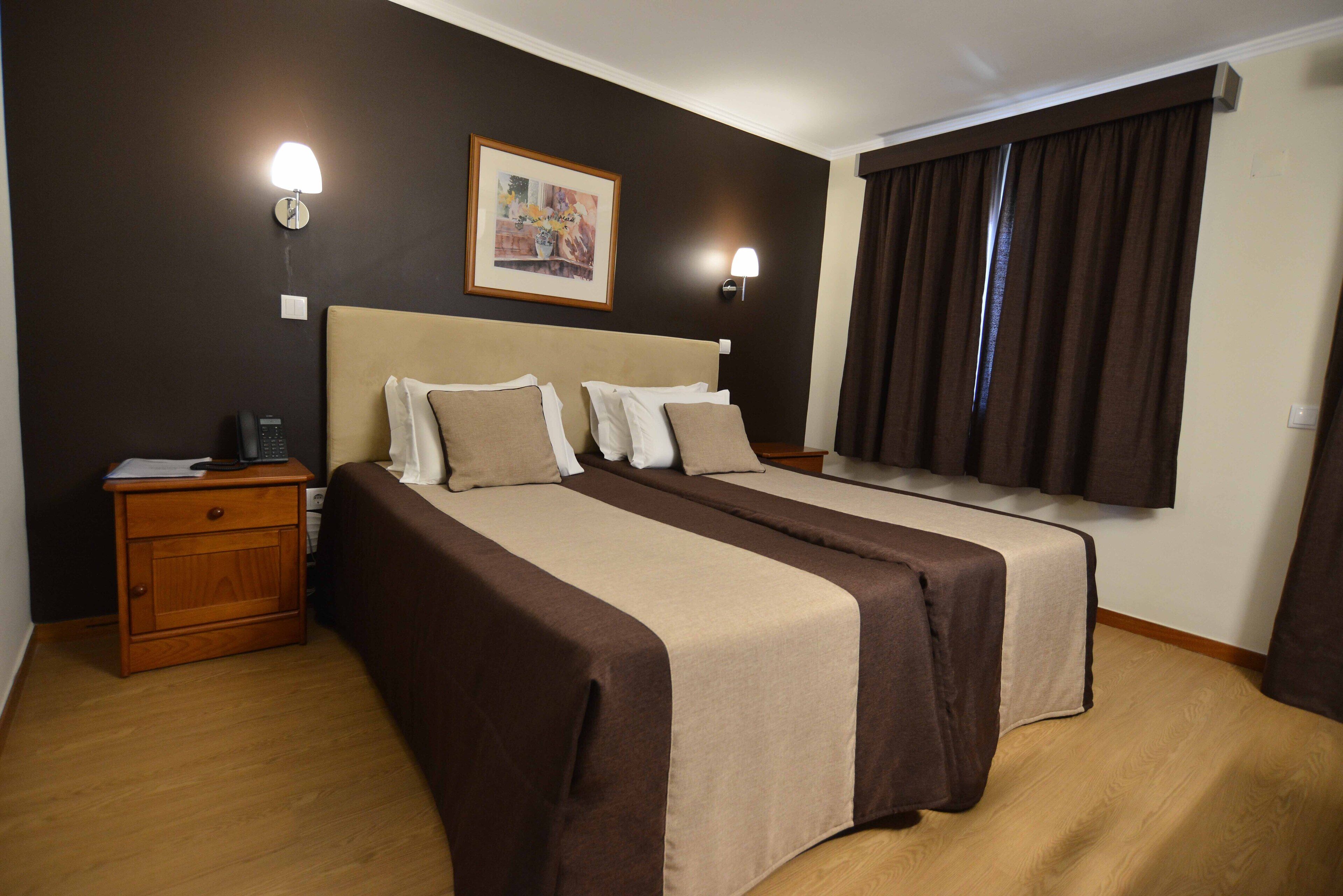 Een hotelkamer met twee bedden, een nachtkastje, een lamp, een schilderij en een raam met gordijnen.