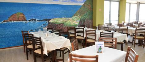 Een restaurant met verschillende tafels en stoelen, een muurschildering van een kustlandschap en grote ramen.