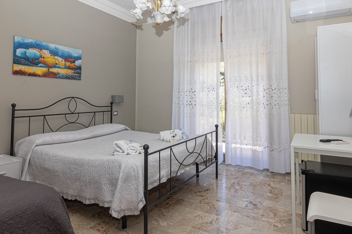 A bedroom with a bed, a desk, a chair, a painting on the wall, and a window with sheer curtains.