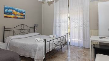 A bedroom with a bed, a desk, a chair, a painting on the wall, and a window with sheer curtains.