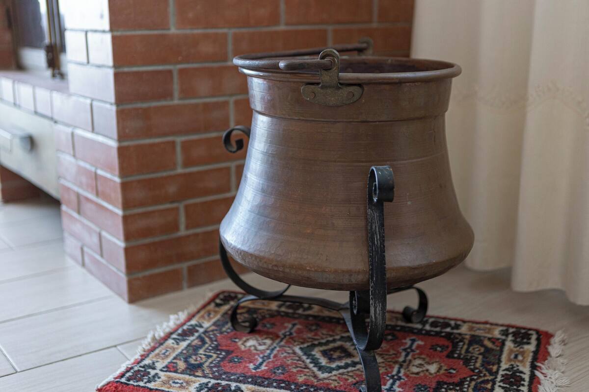 A traditional copper pot on a patterned rug, placed on a tiled floor near a brick wall.