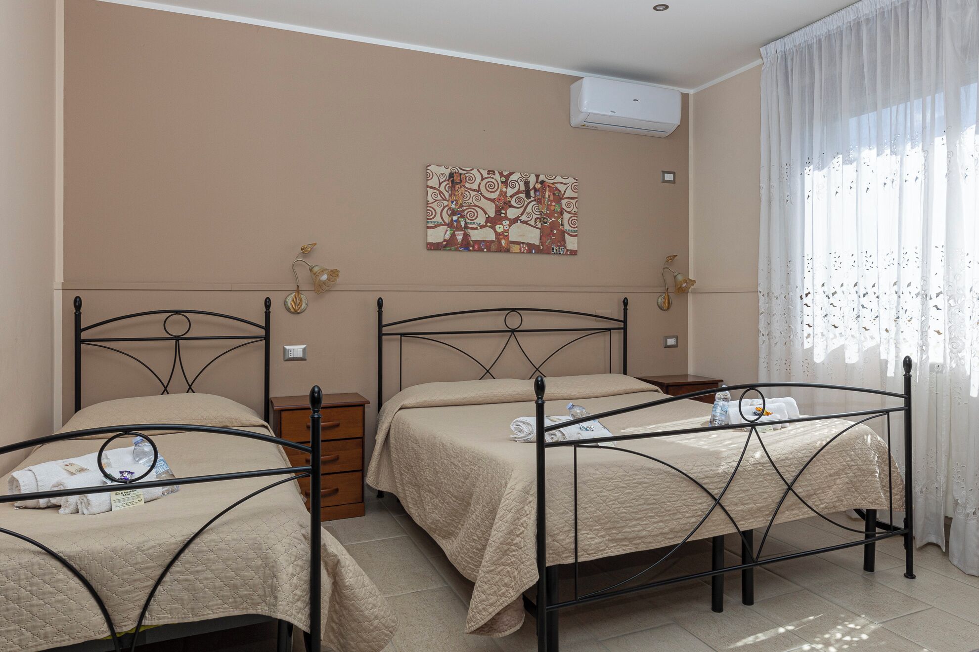 Habitación de hotel con dos camas, una mesita de noche, aire acondicionado instalado en la pared y una ventana con cortinas translúcidas.