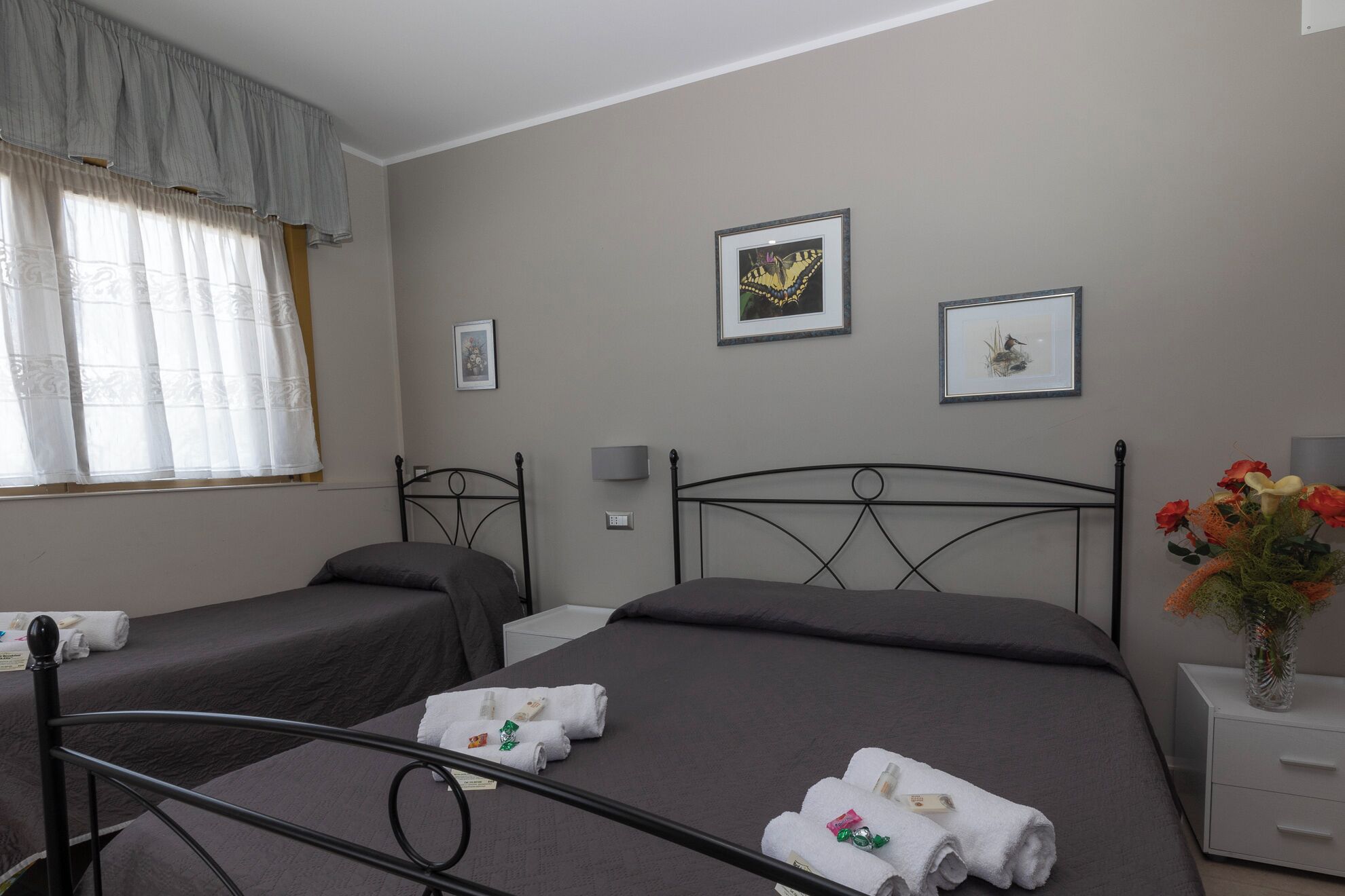 A hotel room with two single beds, a large bed, a window with curtains, a bedside table with a vase of flowers, and wall art.